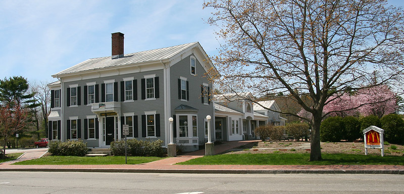 A gray two-story Georgian style historic home with white trim and black shutters sits on a grassy lot. Discreet lettering above the front door reads "McDonalds." A free-standing sign only a few feet tall with the golden arches logo is located to the side of the building in front of evergreen shrubs and redbud trees.